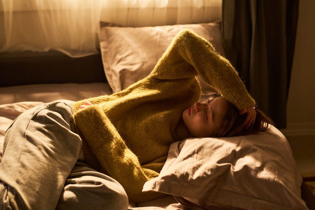 Erschöpfte Frau im gelben Strickpullover liegt im warmen Morgenlicht im Bett – Coaching bei ständiger Müdigkeit in München-Pasing
